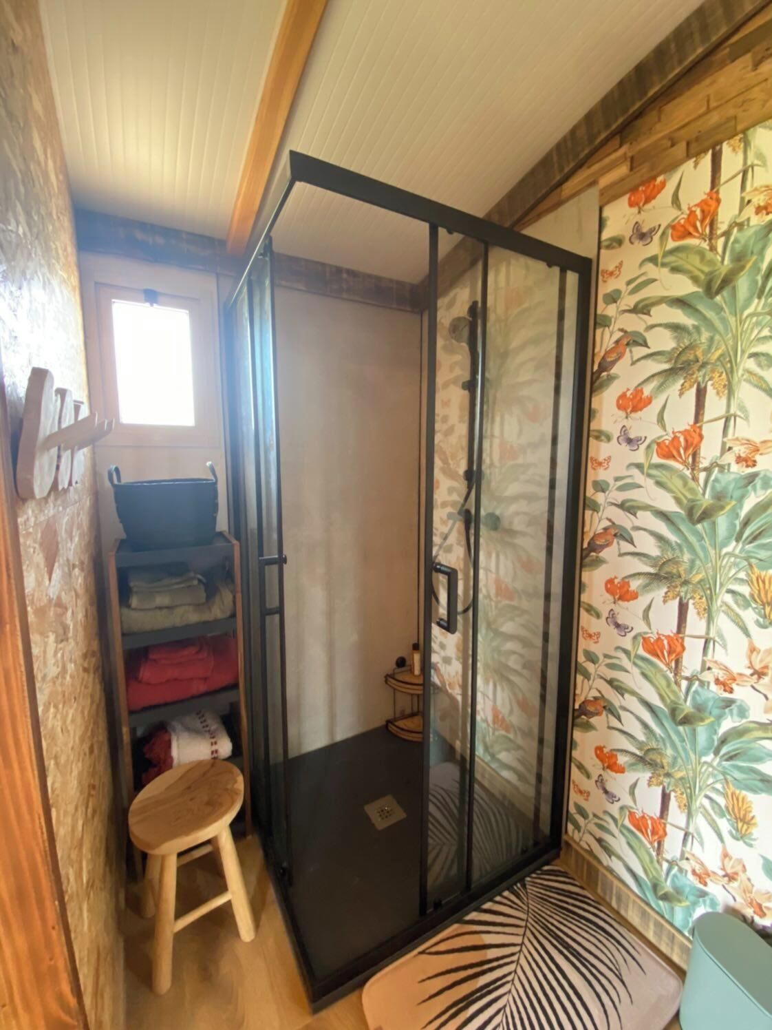 Een kleine badkamer met een douche, een klein krukje en een plank met opgevouwen handdoeken.