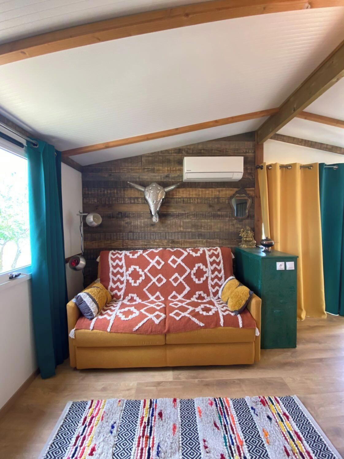 A cozy room with a wooden ceiling, a brown sofa with patterned cushions, a decorative wall-mounted bull's head, and a green cabinet.