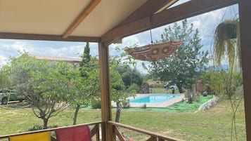 A covered outdoor area with a table and chairs, a hanging plant, and a view of a swimming pool and garden.