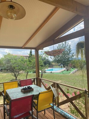 A covered outdoor area with a table and chairs, a hanging plant, and a view of a swimming pool and garden.