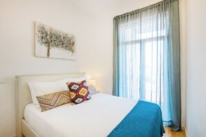 Un lit bien fait, avec une tête de lit blanche, un coussin aux motifs colorés et une couverture bleue. Un tableau représentant un arbre est accroché au mur au-dessus du lit.