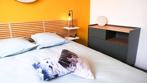 Una habitación de hotel moderna con cabecera de madera, una mesita de noche con lámpara, un espejo redondo y un mueble de almacenamiento empotrado.