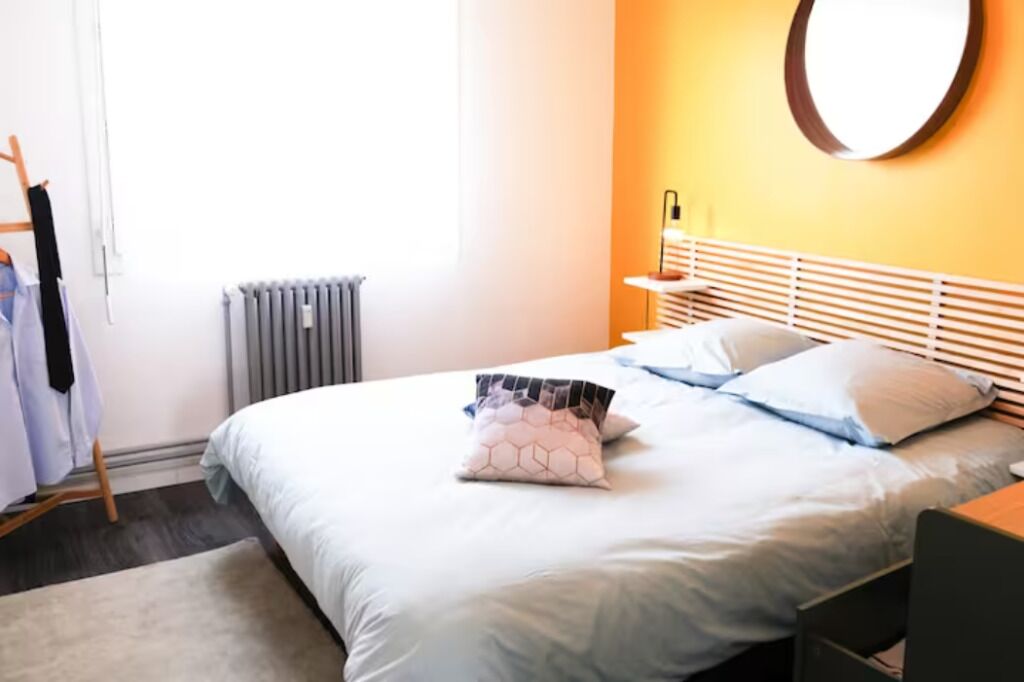 A neatly arranged bedroom with a bed, a bedside table, a chair, a radiator, and a wall-mounted mirror.