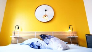 A hotel room with a yellow wall, a round mirror on the wall, a bed with white bedding, and two bedside tables with lamps.