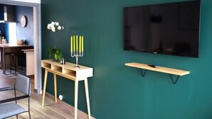 A modern living room with a dark green wall, a flat-screen TV mounted above a wooden shelf, a small table with a potted plant, and a chair.