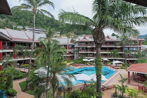 Ein Resort mit einem zentral gelegenen Pool, umgeben von Liegestühlen und Sonnenschirmen, mehrstöckigen Gebäuden mit Balkonen und üppiger Bepflanzung.