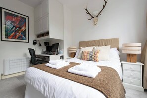 A hotel room with a bed, a desk, a computer, a lamp, and a deer skull on the wall.