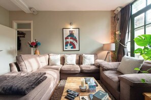 A living room with a sectional sofa, a coffee table with remote controls, a framed artwork, and a large plant.