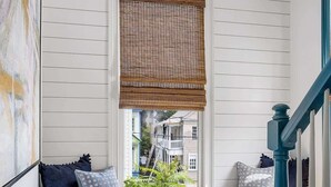 Una habitación con un banco empotrado bajo una ventana con persiana de madera, una escalera con detalles azules y vistas a una casa vecina.