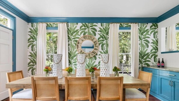 Comedor con mesa y sillas de madera, un espejo grande y un papel tapiz tropical.