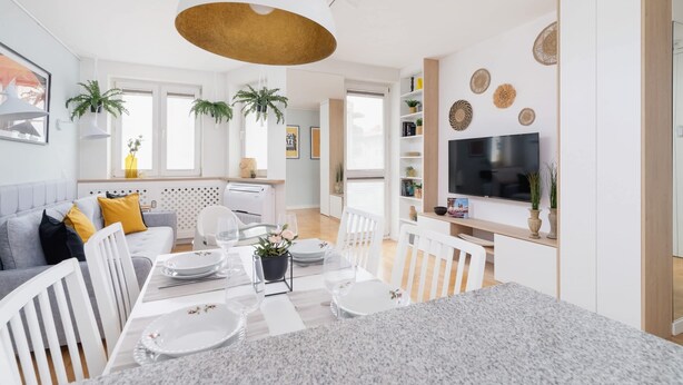 A modern kitchen with a dining area, white furniture, and a TV mounted on the wall.