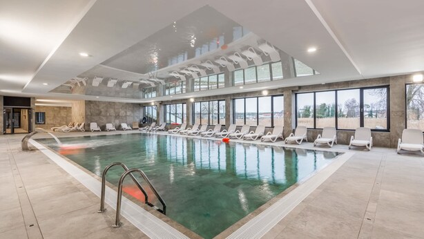 An indoor swimming pool with clear water, surrounded by lounge chairs and large windows offering views of the outdoors.