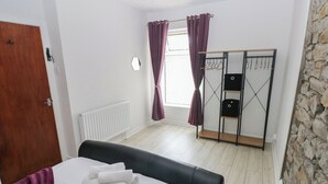 A bedroom with a bed, a wooden door, a radiator, a window with purple curtains, a wardrobe, and a stone wall. - Brynawel (Burry Port)