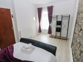 A bedroom with a bed, a wooden door, a radiator, a window with purple curtains, a wardrobe, and a stone wall.