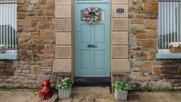 A stone building with a teal door, a number plaque, and flower baskets on the steps. - Brynawel (Burry Port)