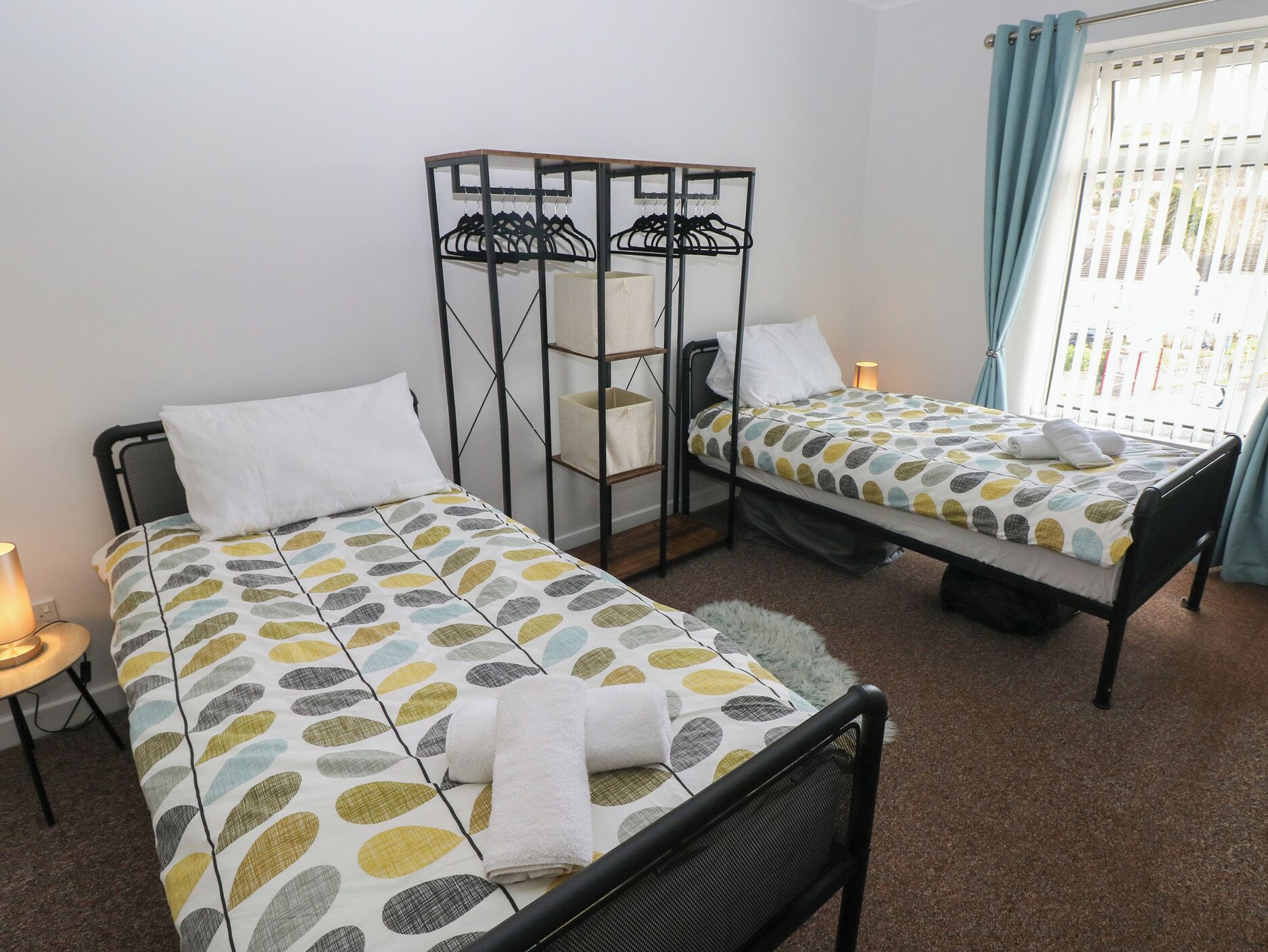A bedroom with two single beds, a metal wardrobe, a small table, and a window with curtains.