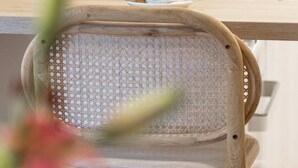 Una silla de madera con respaldo tejido, una piña sobre la mesa, y una taza blanca y una cafetera al fondo.