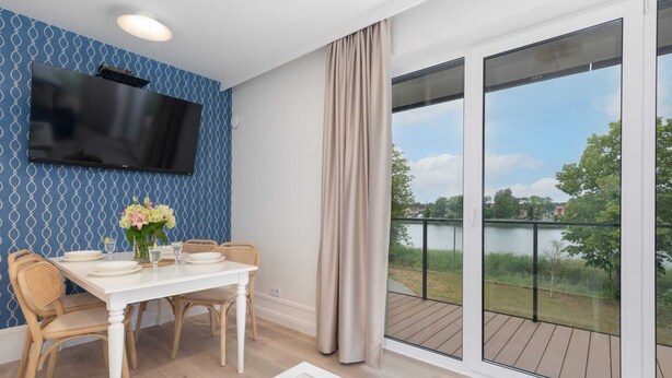 A dining area with a table set for four, a flat-screen TV mounted on the wall, and a view of a lake through large windows. - Apartment with 2 Bedrooms | Parking, SPA, Close to the Beach (Dziwnów)