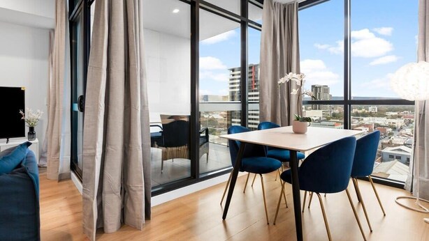 A modern dining area with a table and chairs, large windows with curtains, and a view of the cityscape.
