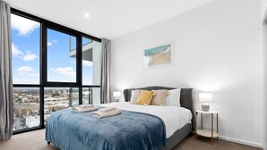 A modern bedroom with a large bed, a view of the city, and a small bedside table with a lamp.