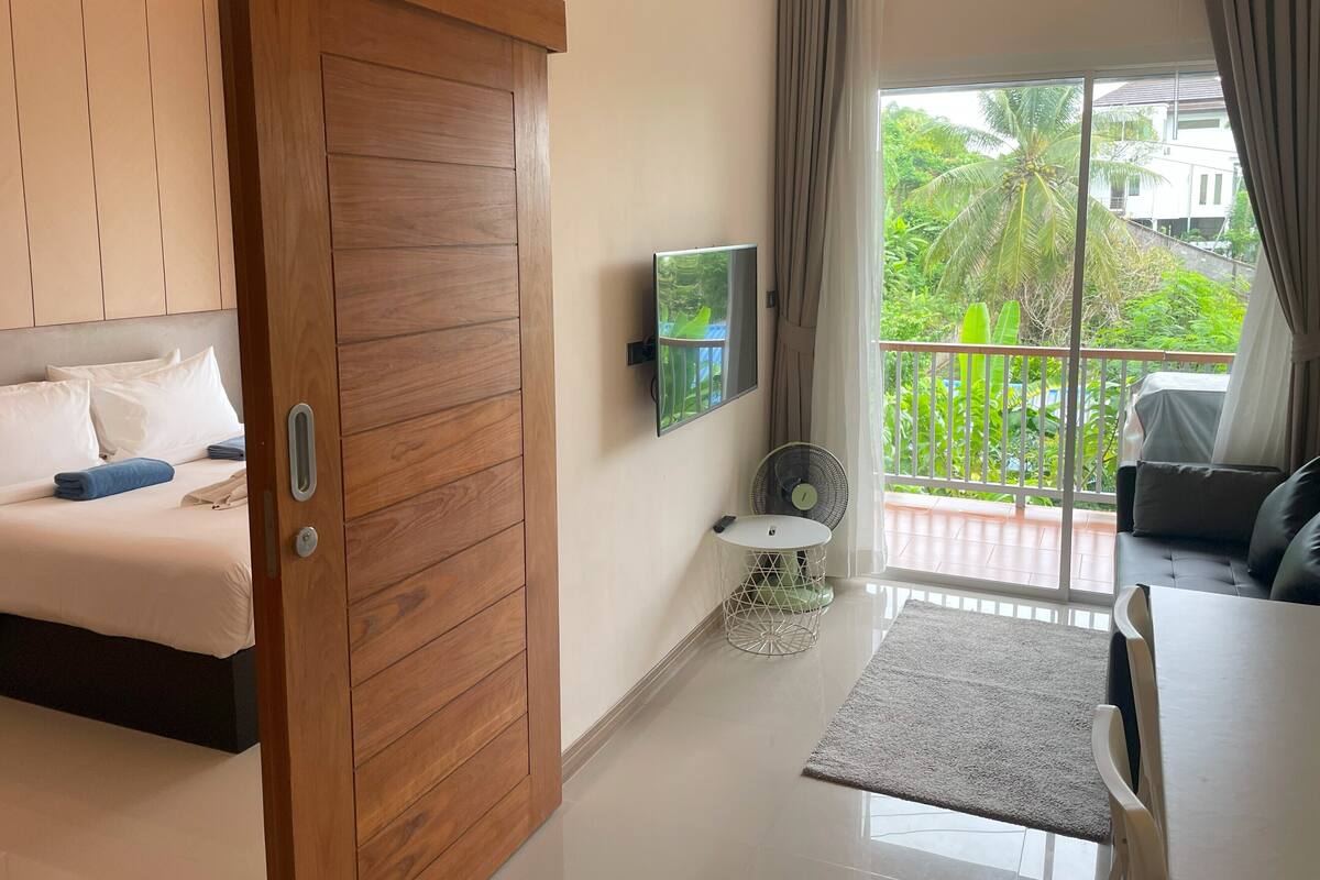 A modern hotel room with a wooden wardrobe, a bed with white linens, a TV mounted on the wall, a small table with a fan, and a balcony with a view of greenery.