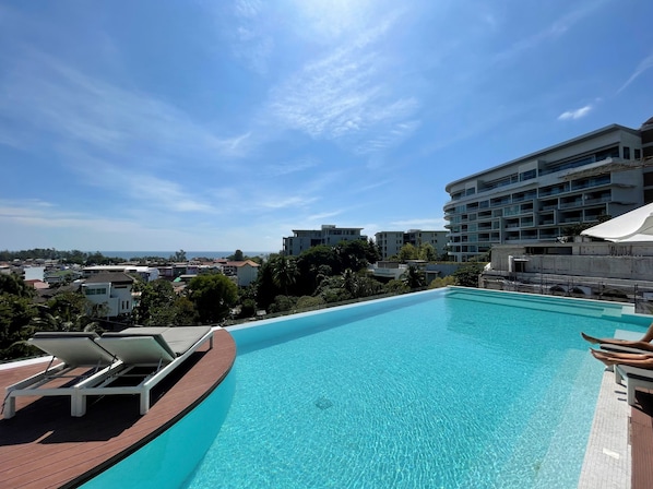 A clear blue pool with lounge chairs, surrounded by buildings and trees, under a sunny sky.