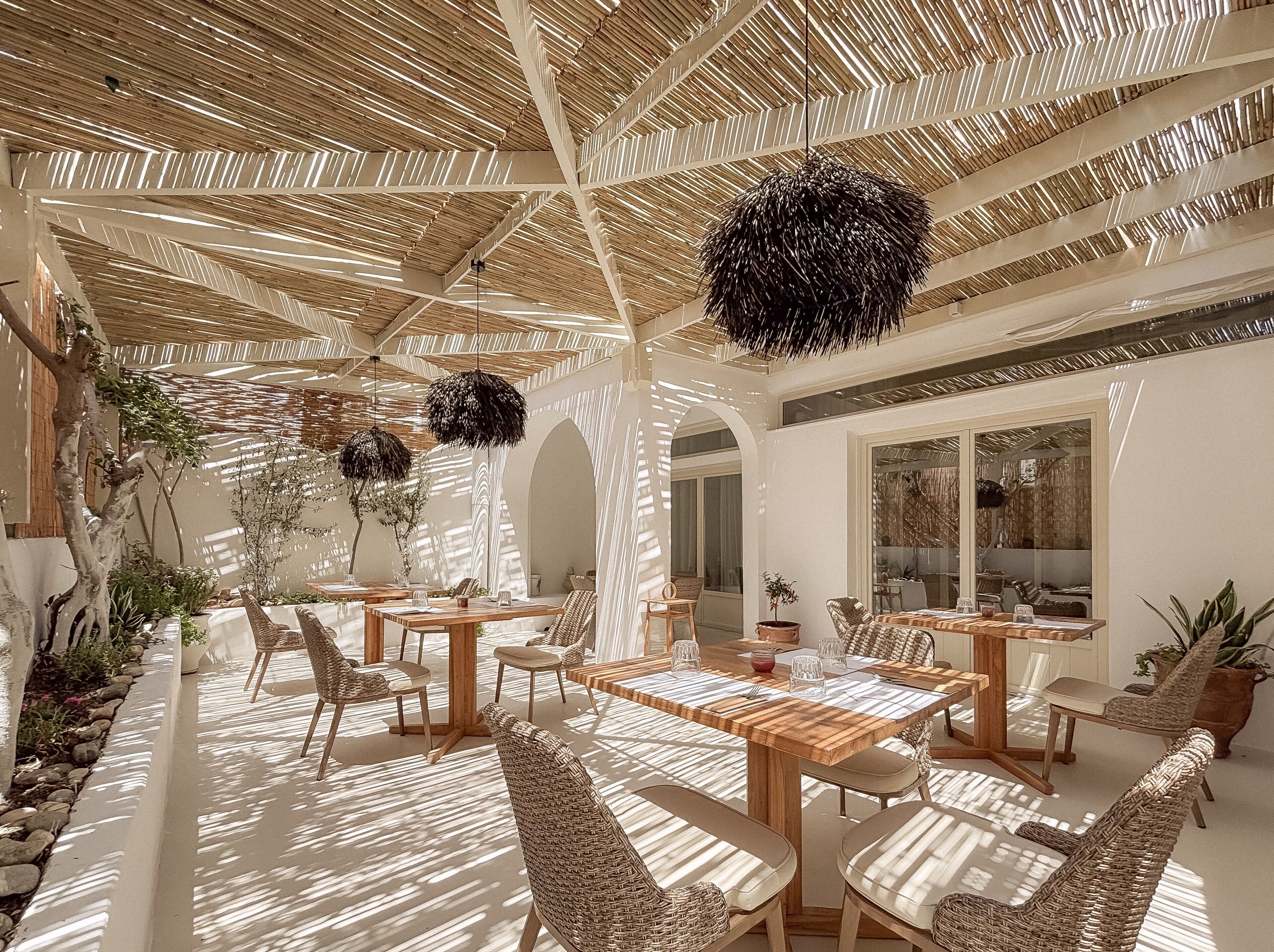 Un amplio espacio exterior con mesas de madera y sillas de mimbre, techo de paja y luz natural que se filtra a través de él.