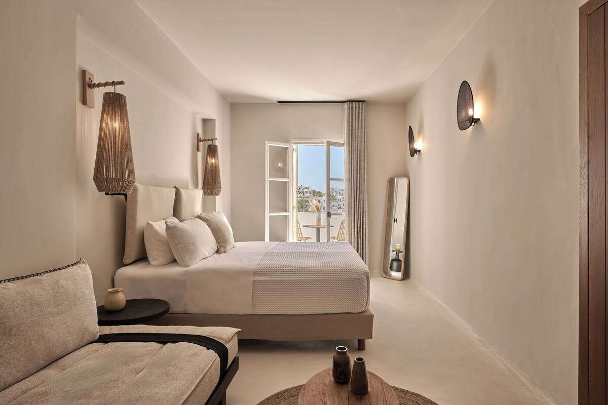 Une chambre moderne avec un grand lit, un canapé, une petite table ornée de vases et une vue sur l’extérieur grâce à une porte ouverte.
