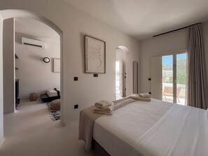 A bedroom with a large bed, a sitting area with a sofa, a dining table, and a view of the outdoors. - Utopia Hotel (Koufonisia)