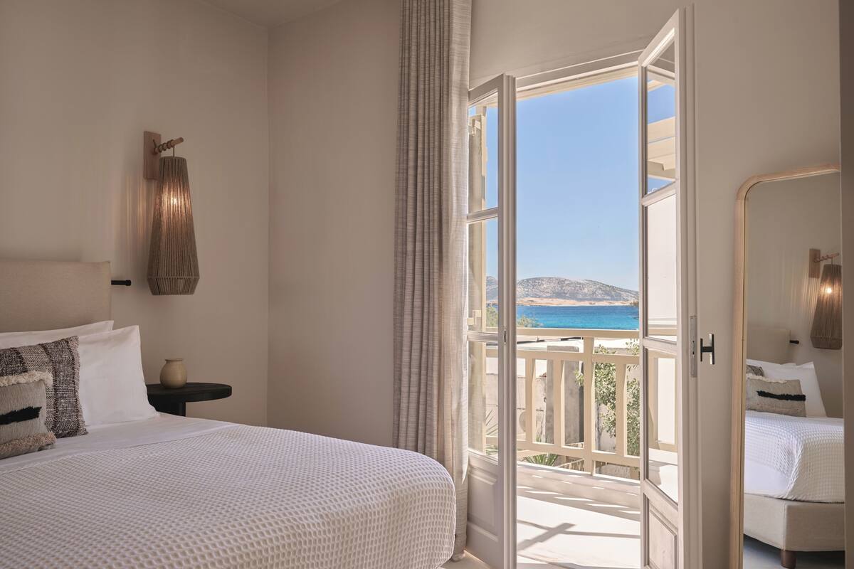 Une chambre à coucher avec un grand lit, une table de chevet, une lampe et une vue sur la mer, visible par une porte ouverte.