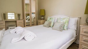 Een opgemaakt bed met witte lakens, een spiegel, een kledingkast en een nachtkastje met een lamp.