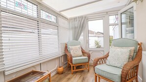 Een zonnige kamer met rieten stoelen, een tafeltje met kinderspeelgoed en uitzicht door een raam met luiken.