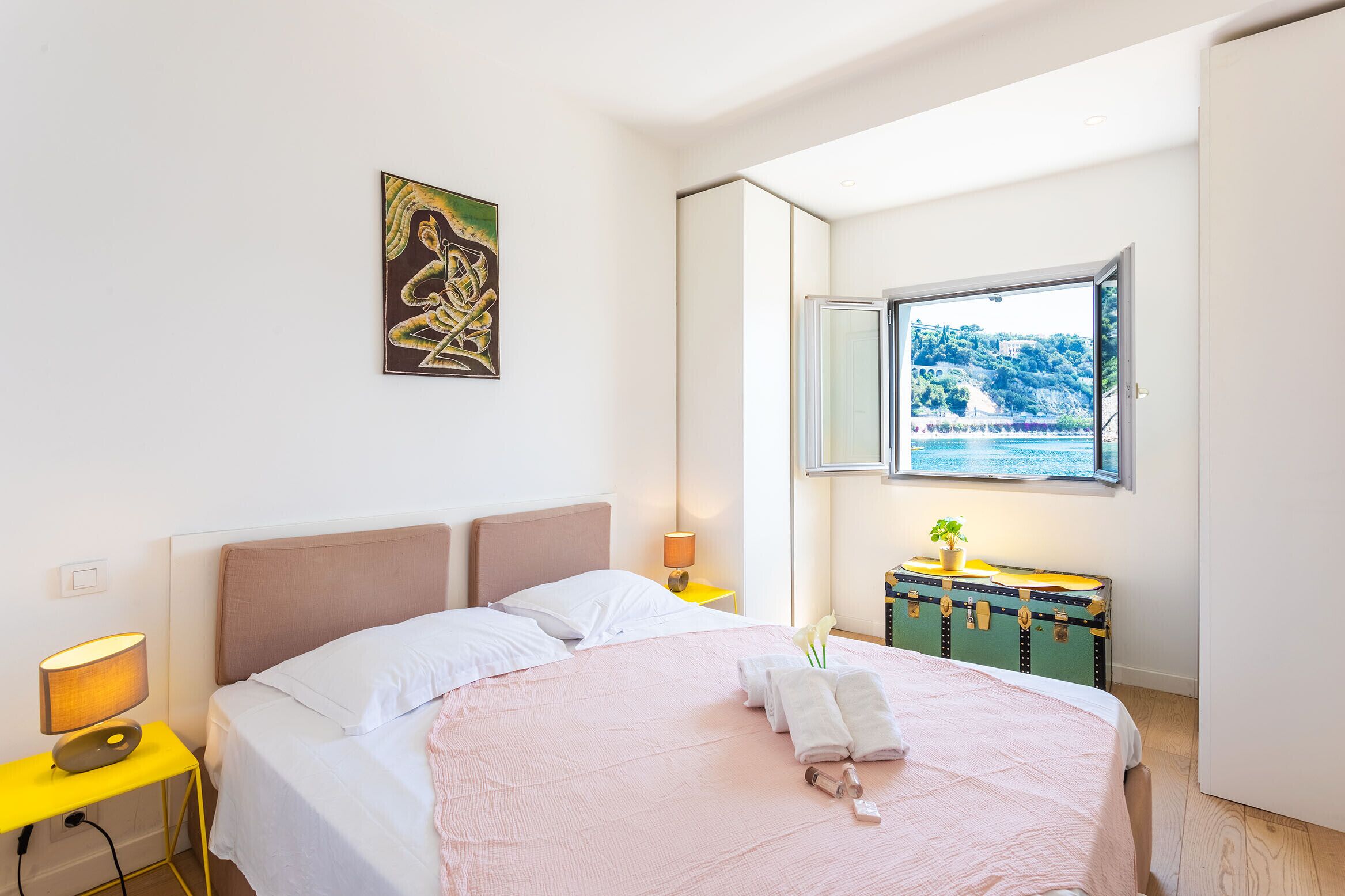 A bedroom with a bed, a bedside table with a lamp, a window with a view, and a wall-mounted painting.