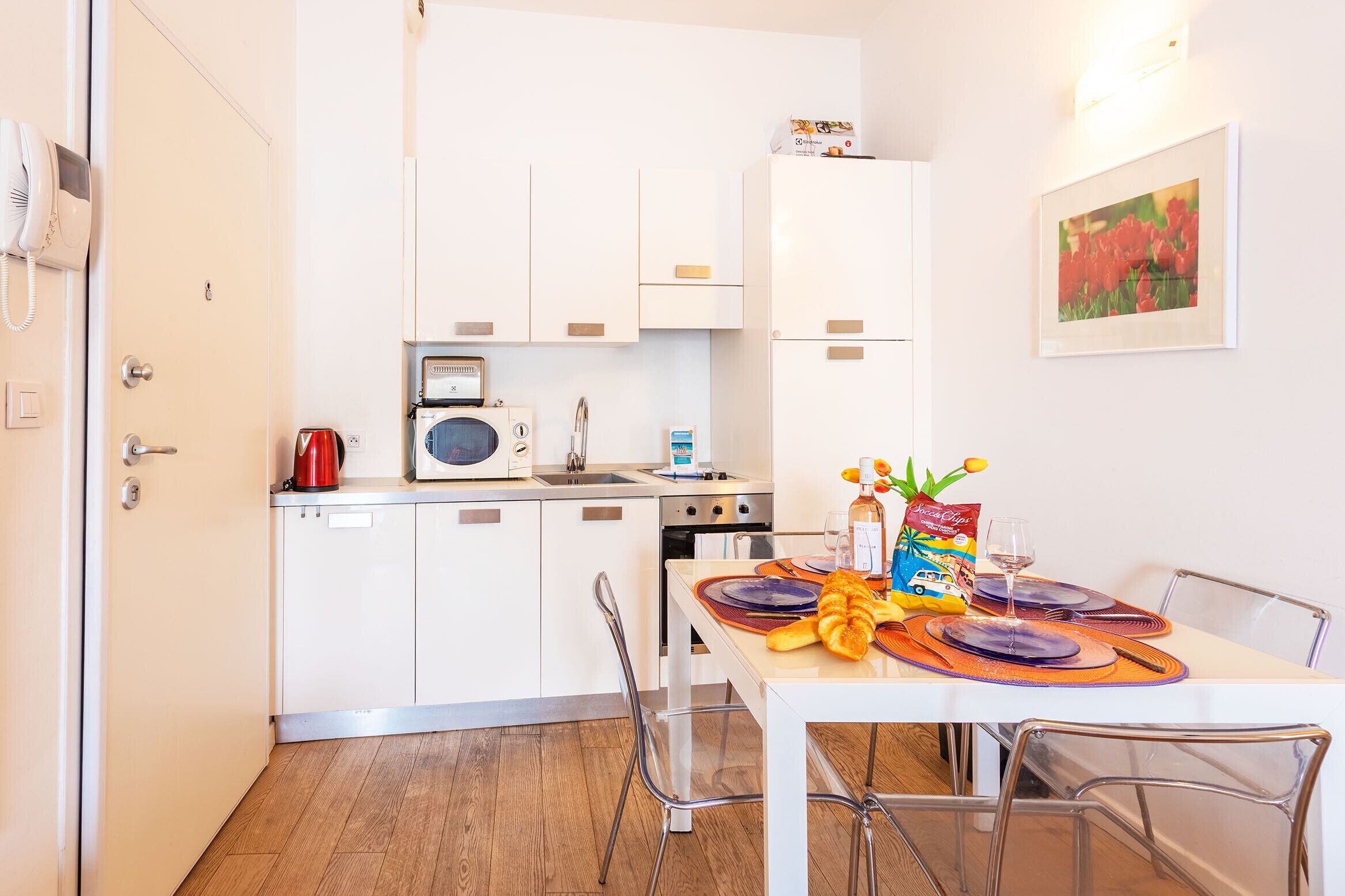 A compact kitchen with white cabinets, a red kettle, and a microwave. A dining table with orange placemats and a bottle of water is set.