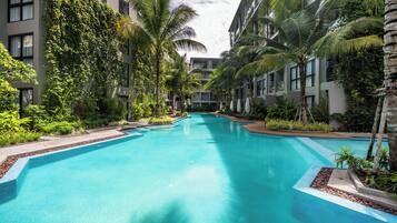 A swimming pool surrounded by multi-story buildings with balconies, lush greenery, and a clear blue sky.