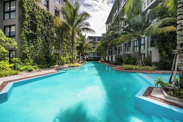 A swimming pool surrounded by multi-story buildings with balconies, lush greenery, and a clear blue sky.