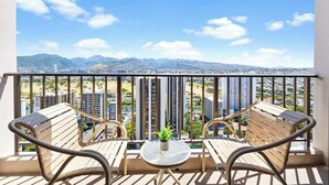 A balcony with two wooden chairs, a small table, and a potted plant. - Waikiki Sunset 28F w/ pool, views & beach walk (Honolulu)