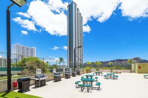 A rooftop patio with barbecue grills, picnic tables, and a view of a tall building and mountains. - Waikiki Sunset 28F w/ pool, views & beach walk (Honolulu)