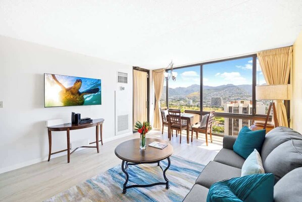 A modern living room with a large window, a flat-screen TV, a round coffee table, and a sofa with cushions. - Waikiki Sunset 28F w/ pool, views & beach walk (Honolulu)