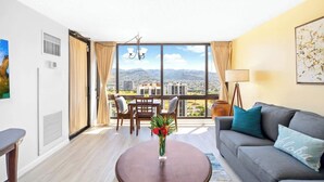 A modern living room with a sofa, coffee table, and dining table, large windows with a city view, and a balcony. - Waikiki Sunset 28F w/ pool, views & beach walk (Honolulu)