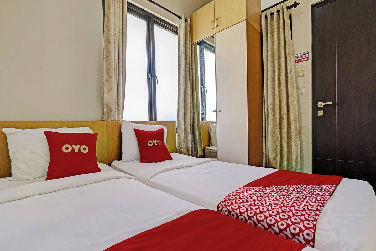 A hotel room with two beds, each with white bedding and red pillows. There is a large window with curtains, a wardrobe, and a door with a 'No Smoking' sign.