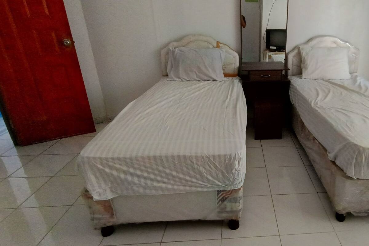 Ein Zimmer mit zwei Einzelbetten, einem kleinen Holfnachtkästchen, einer roten Tür und einem weiß gefliesten Boden.