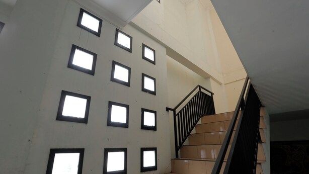 Una escalera con barandilla negra y escalones color beige, que lleva a un rellano con varias ventanas cuadradas.