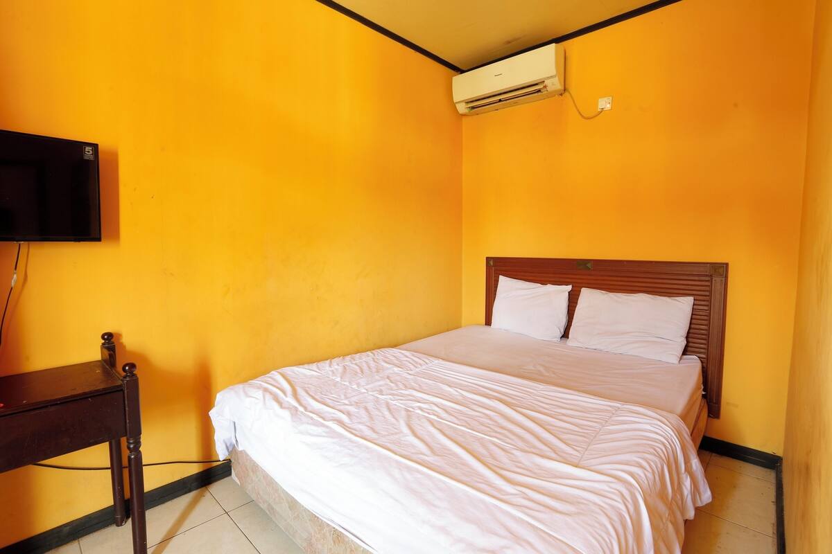 A room with a bed, a wooden nightstand, a flat-screen TV mounted on the wall, and an air conditioning unit.