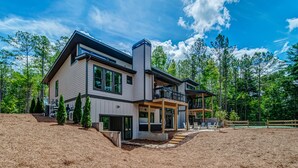 A modern two-story house with a large deck, surrounded by a wooded area. - Amazing Grace ~ Indoor Heated Pool ~ Pickleball ~ Basketball ~ Hot Tub ~ Fire Pit (Ellijay)