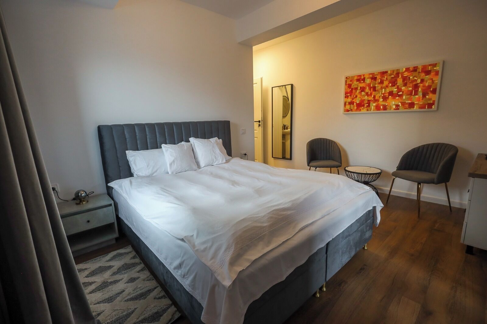 A modern hotel room with a large bed, a bedside table, a chair, a small round table, and a mirror.
