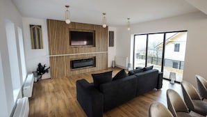 A modern living room with a black sectional sofa, a flat-screen TV mounted on a wooden panel wall, and a large window offering a view of the outside.