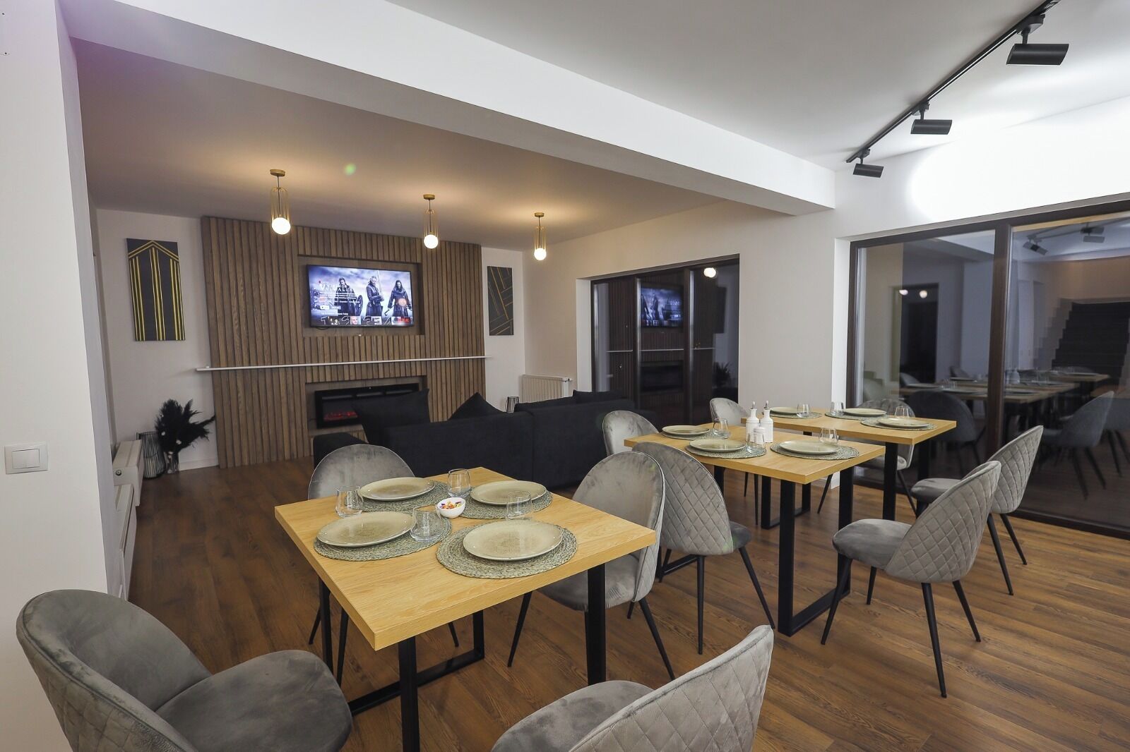 Een moderne eetruimte met houten tafels, grijs gestoffeerde stoelen en een flatscreen televisie aan de muur.