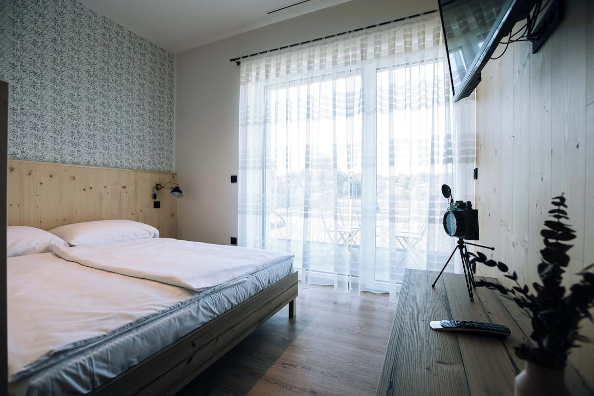 A bedroom with a wooden bed, a TV mounted on the wall, and a balcony with a view of the city.