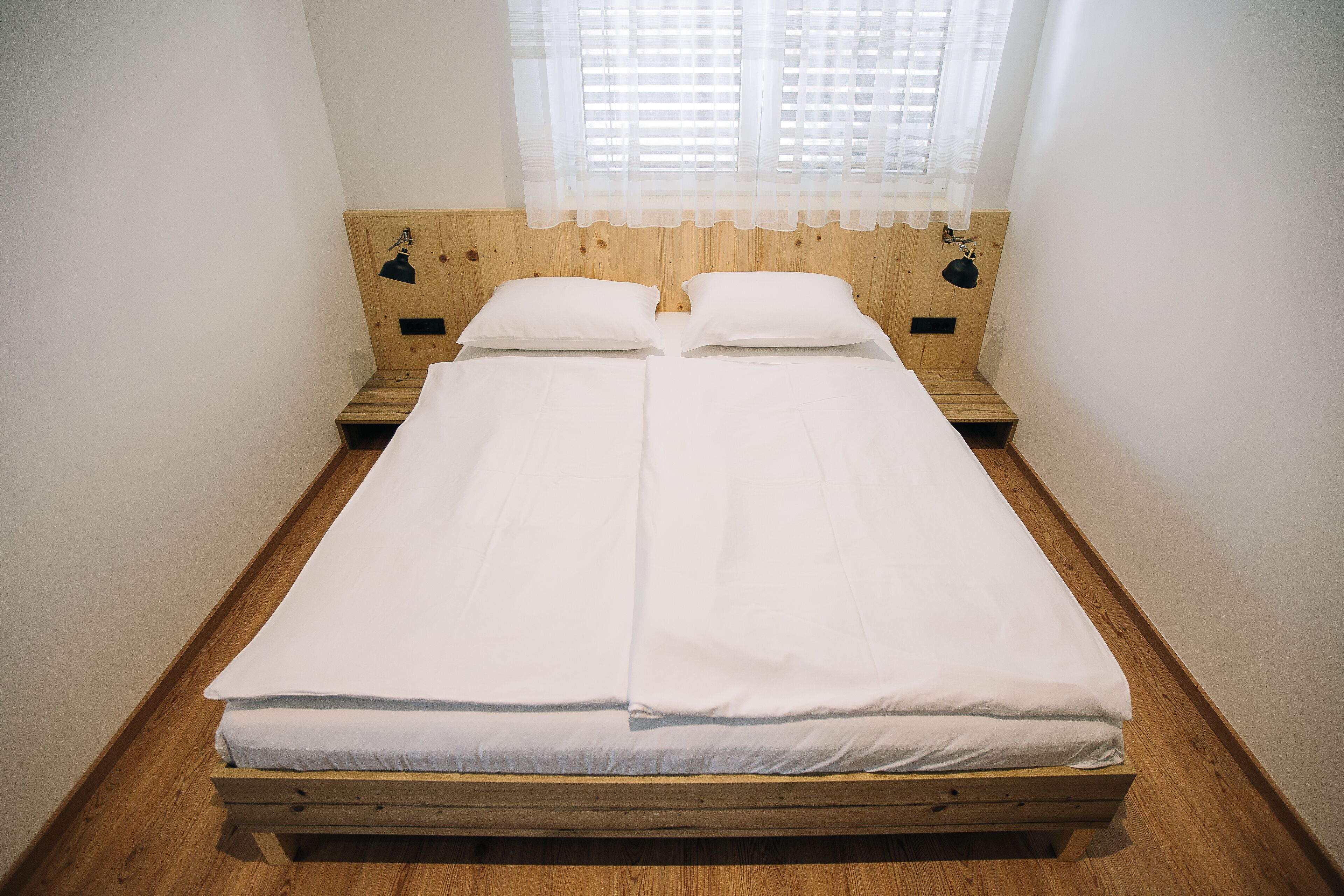 A double bed with a wooden headboard and two bedside tables, a window with sheer curtains, and a wooden floor.